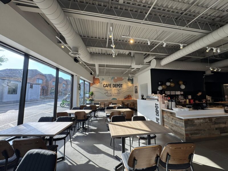 Cafe Depot Interior A bright, modern interior of Café Dépôt in Downtown Milton, featuring large windows with sunlight streaming in, wooden tables and chairs neatly arranged, and a cozy mural wall at the back with “Café Dépôt” signage. Industrial ceiling pipes and a view of the quiet street outside complete the relaxed vibe.