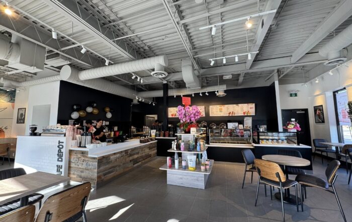 Interior of Café Dépôt showing the café counter with staff preparing drinks. The setup includes rustic wood paneling, a display case filled with desserts, pink orchids at the counter, and a black menu board on the back wall. The space is airy and well-lit with a mix of seating arrangements and natural light from tall windows.